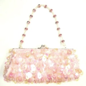 Peut inclure: Un sac à main rose clair avec une poignée perlée. Le sac est recouvert de petites paillettes irisées. La poignée est faite de perles blanches avec des accents roses et rouges. Le fermoir est argenté.