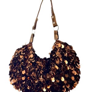 May include: A brown sequined handbag with a matching strap. The bag is crescent-shaped and covered in small, round sequins. The strap is a darker brown and attached to the bag with metal rings.