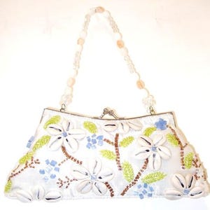 May include: White satin clutch bag with floral and seashell embellishments. The bag features white flowers, green leaves, and blue accents. The handle is made of clear and peach-coloured beads. A silver clasp secures the top.