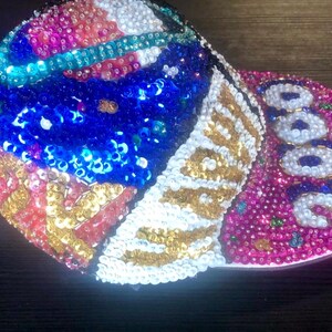 Op de afbeelding: Een met pailletten versierde baseballcap met een levendig ontwerp. De cap heeft een mix van kleuren, waaronder rode, witte, blauwe en gouden pailletten. De klep is roze met het nummer "2000" in witte pailletten. Een stijlvol accessoire.