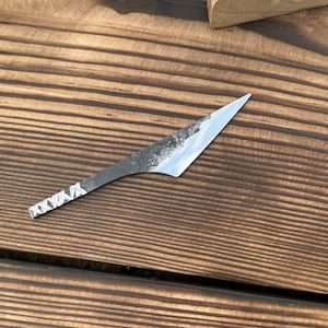 Könnte beinhalten: Eine kleine, spitze Metallklinge mit einem dunklen, texturierten Finish nahe dem Rücken und einer polierten, reflektierenden Schneide. Der Griffbereich weist eine Reihe sich wiederholender, geätzter X-Muster auf und endet in einer scharfen Spitze.
