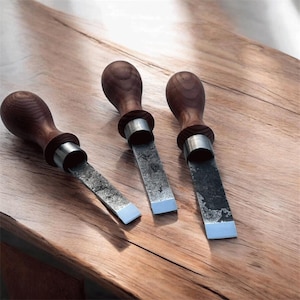 May include: Three woodworking chisels with dark wooden handles and silver metal collars. The blades are rectangular with a light blue edge. The tools are arranged on a wooden surface.