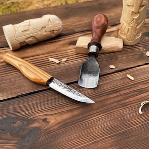May include: A wood carving set featuring a fixed-blade knife with a light-colored wooden handle, a metal gouge with a dark wooden handle, and two carved wooden figures. The tools have textured metal blades and are arranged on a dark wood surface.