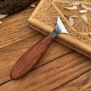 May include: A wood carving knife with a dark wooden handle and a silver blade. The knife is resting on a wooden surface next to a carved wooden block. Wood shavings are scattered around the knife and block.