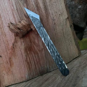 Puede incluir: Un cuchillo artesanal con un mango gris oscuro texturizado y una hoja plateada. El cuchillo descansa sobre un trozo de madera con vetas naturales. La hoja está en ángulo y el cuchillo parece afilado.