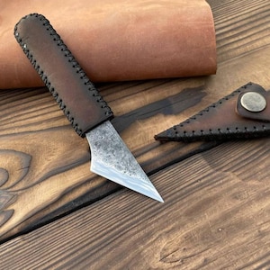 May include: A fixed-blade knife with a dark brown leather-wrapped handle featuring visible stitching along the edges. The blade has a textured, hammered metal finish and a sharp, angled point. A matching leather sheath with a brass snap button.