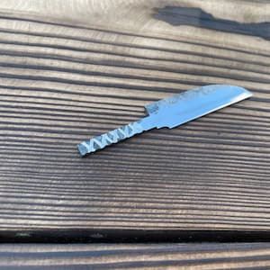 May include: A small, single-piece metal knife with a polished, curved blade and a textured, dark grey handle featuring a repeating cross-hatch pattern. The blade shows a subtle, mottled finish near the spine, transitioning to a smooth edge.