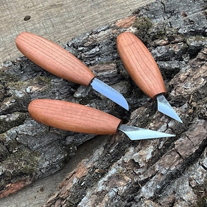 May include: Three wood carving knives with wooden handles and silver blades are arranged on a textured, bark-covered surface. The knives have a curved blade design, suitable for detailed woodworking projects.
