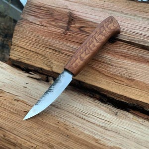 Op de afbeelding: Een handgemaakt mes met een houten handvat en een zilveren lemmet. Het handvat heeft een getextureerde, lichtbruine afwerking. Het lemmet is scherp en puntig, met een enigszins ruwe rand. Het mes ligt op een houten oppervlak.
