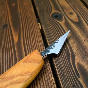 Może przedstawiać: Zbliżenie noża do rzeźbienia z drewnianą rękojeścią i srebrnym ostrzem. Ostrze ma ostry czubek i ciemny, teksturowany obszar w pobliżu rękojeści. Rękojeść jest w jasnobrązowym kolorze. Nóż jest ustawiony na tle ciemnego drewna.