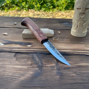 Peut inclure: Un couteau à sculpter à lame fixe doté d'une lame en acier poli et d'un manche en bois brun foncé profilé. Le couteau repose sur un petit bloc de bois clair, placé à côté d'une figurine en bois sculpté sur une surface en bois rustique.