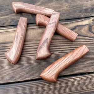 May include: Four smooth, polished wooden knife handles with a distinct reddish-brown grain pattern. Each handle features a curved, ergonomic shape designed for a secure grip, showcasing natural wood textures and variations in color throughout.