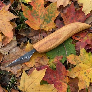 Puede incluir: Un cuchillo artesanal con mango de madera y hoja oscura y texturizada descansa sobre un lecho de coloridas hojas de otoño. Las hojas muestran tonos de amarillo, naranja y rojo.