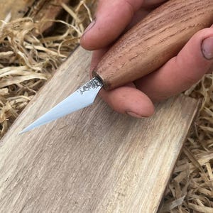 Könnte beinhalten: Ein Holzschnitzmesser mit einem hellbraunen Holzgriff und einer silbernen Klinge. Das Messer wird über einem Stück hellbraunem Holz gehalten. Die Klinge ist scharf und spitz, für detaillierte Schnitzarbeiten konzipiert.