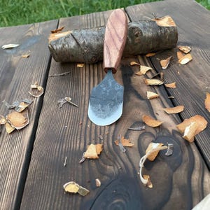 Könnte beinhalten: Ein handgeschmiedetes Metallmesser mit einer breiten, abgerundeten Klinge und einer dunklen, strukturierten Oberfläche. Der Griff ist aus poliertem braunem Holz mit sichtbarer Maserung gefertigt. Das Messer steht vertikal an einem Holzscheit.