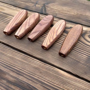 Puede incluir: Cinco mangos de madera alargados en forma de lágrima con un acabado suave y pulido y patrones de grano natural visibles. Cada mango presenta una parte superior redondeada y una base plana y cónica con un pequeño orificio circular.