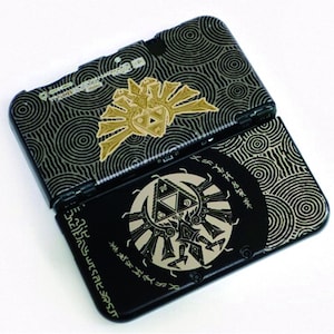 Könnte beinhalten: Eine schwarze tragbare Spielkonsole mit einem goldenen Triforce-Emblem und komplizierten kreisförmigen Linienmustern. Das Gerät zeigt stilisierte Hylian-Schriftzeichen und eine kreisförmige Grafik mit einem zentralen Dreieckssymbol.
