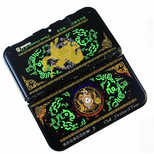 Op de afbeelding: Een zwarte draagbare spelcomputer met gouden en neongroene grafische ontwerpen. Het bovenpaneel toont een gouden paard-silhouet, terwijl het onderpaneel een gedetailleerde klok bevat. De tekst Classic Ocarina of Time staat op beide panelen vermeld.
