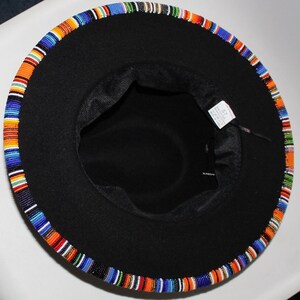 May include: Black fedora hat with a colorful beaded band around the brim. The band features a pattern of blue, orange, green, and white beads. The hat has a black interior and a tag with the number 57.
