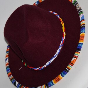 May include: A burgundy felt hat with a wide brim. The brim is decorated with a colorful beaded trim in shades of orange, blue, and white. A smaller beaded rope in red, white, and yellow accents the hat.