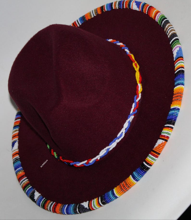 May include: A burgundy felt hat with a wide brim. The brim is decorated with a colorful beaded trim in shades of orange, blue, and white. A smaller beaded rope in red, white, and yellow accents the hat.