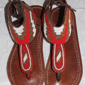 Op de afbeelding: Bruin leren sandalen met ingewikkeld kralenwerk in rood, wit en goud. De sandalen hebben een teenslipper-ontwerp met een gestikte zool en bruine veters.