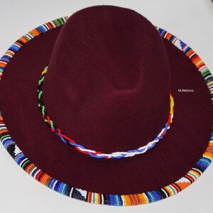 May include: A burgundy fedora-style hat with a wide brim. The brim is adorned with a colorful beaded trim in shades of blue, orange, and white. A beaded band in similar colors encircles the crown. The text "MJ&Sons" is visible on the hat.