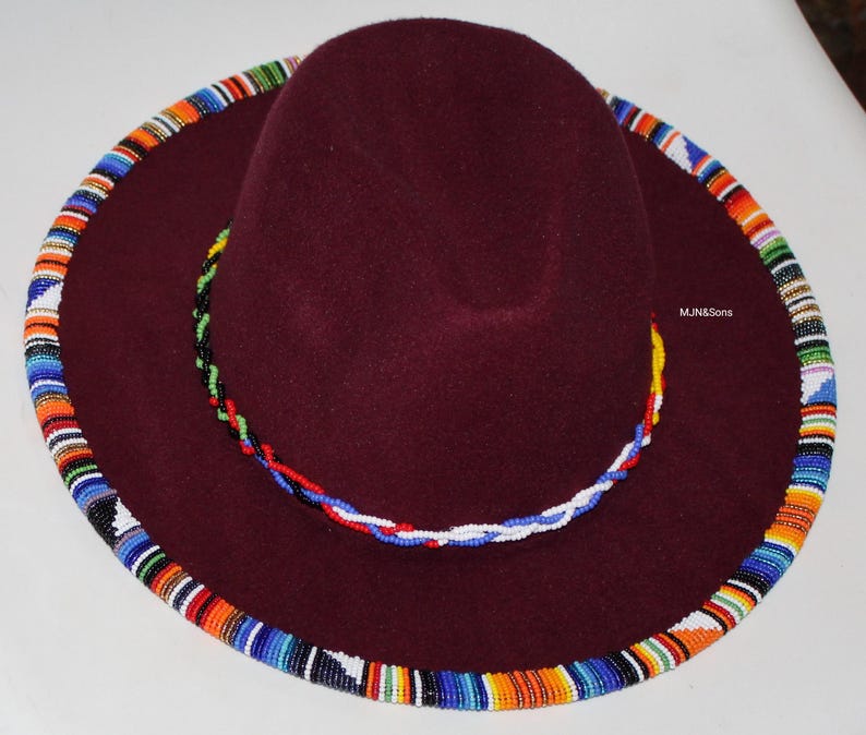 May include: A burgundy fedora-style hat with a wide brim. The brim is adorned with a colorful beaded trim in shades of blue, orange, and white. A beaded band in similar colors encircles the crown. The text "MJ&Sons" is visible on the hat.