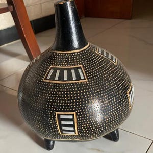 Op de afbeelding: Een zwarte decoratieve vaas met een bolvormige body en een smalle hals. De vaas heeft een textuur met kleine stippen en geometrische patronen in wit en beige. Hij staat op drie kleine voetjes.