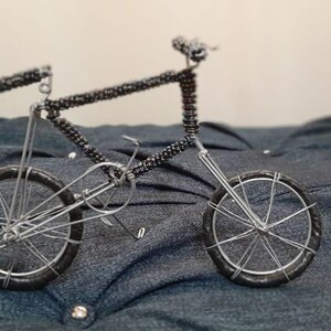 Puede incluir: Una escultura de bicicleta en miniatura hecha de alambre plateado. El cuadro y el manillar están detallados con pequeñas cuentas oscuras. Las ruedas tienen neumáticos negros y radios plateados. La pieza decorativa se encuentra sobre una superficie azul oscuro acolchada.