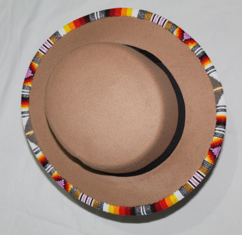 Puede incluir: Un sombrero fedora color canela con un ribete de cuentas. El ribete presenta un patr&oacute;n de cuentas rojas, naranjas, amarillas, blancas, grises y moradas. El sombrero tiene una banda interior negra y una corona redondeada.