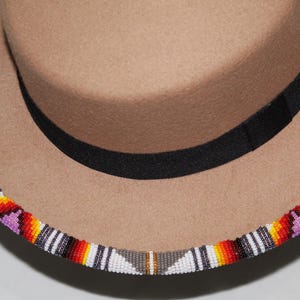 Puede incluir: Un sombrero fedora color canela con una banda negra y un ribete de cuentas. El ribete presenta un patr&oacute;n colorido de cuentas rojas, naranjas, amarillas, blancas y moradas. Un accesorio elegante.