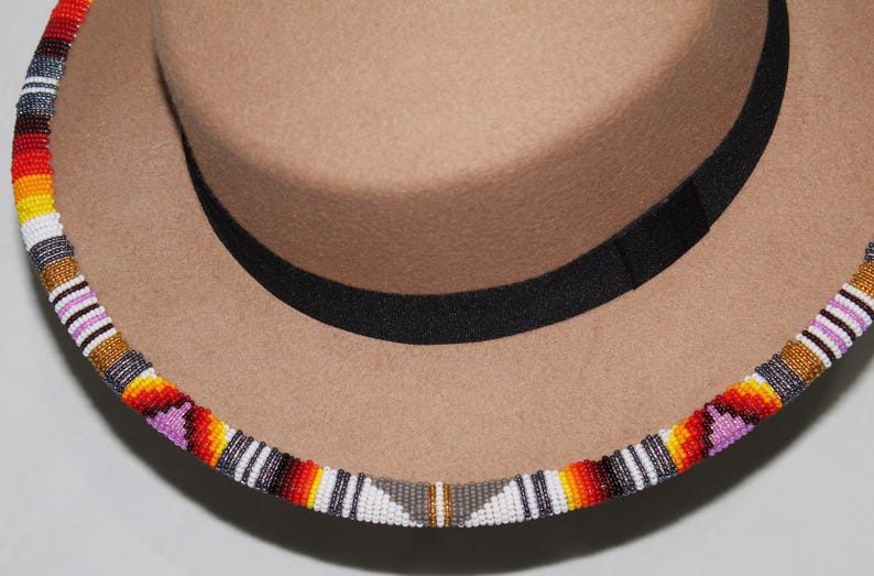 Puede incluir: Un sombrero fedora color canela con una banda negra y un ribete de cuentas. El ribete presenta un patr&oacute;n colorido de cuentas rojas, naranjas, amarillas, blancas y moradas. Un accesorio elegante.