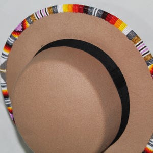 Puede incluir: Un sombrero color canela con una banda negra y un borde con cuentas. El borde est&aacute; decorado con un patr&oacute;n colorido de cuentas blancas, grises, amarillas, naranjas, rojas y moradas. El sombrero tiene una forma de fedora cl&aacute;sica.