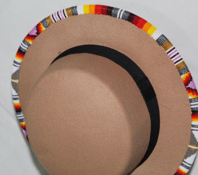 Puede incluir: Un sombrero color canela con una banda negra y un borde con cuentas. El borde est&aacute; decorado con un patr&oacute;n colorido de cuentas blancas, grises, amarillas, naranjas, rojas y moradas. El sombrero tiene una forma de fedora cl&aacute;sica.