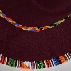 May include: A maroon hat with a beaded trim in a vibrant pattern of orange, white, blue, and green. A smaller beaded strand in yellow, blue, and orange is also visible. The hat's brim is decorated with colorful beads.