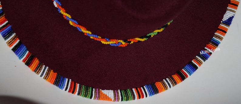 May include: A maroon hat with a beaded trim in a vibrant pattern of orange, white, blue, and green. A smaller beaded strand in yellow, blue, and orange is also visible. The hat's brim is decorated with colorful beads.