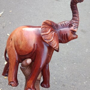 May include: A handcrafted wooden elephant sculpture with a polished, reddish-brown finish. The elephant has detailed carvings on its ears, trunk, and body, and is posed with its trunk raised. This decorative piece is a unique home decor item.