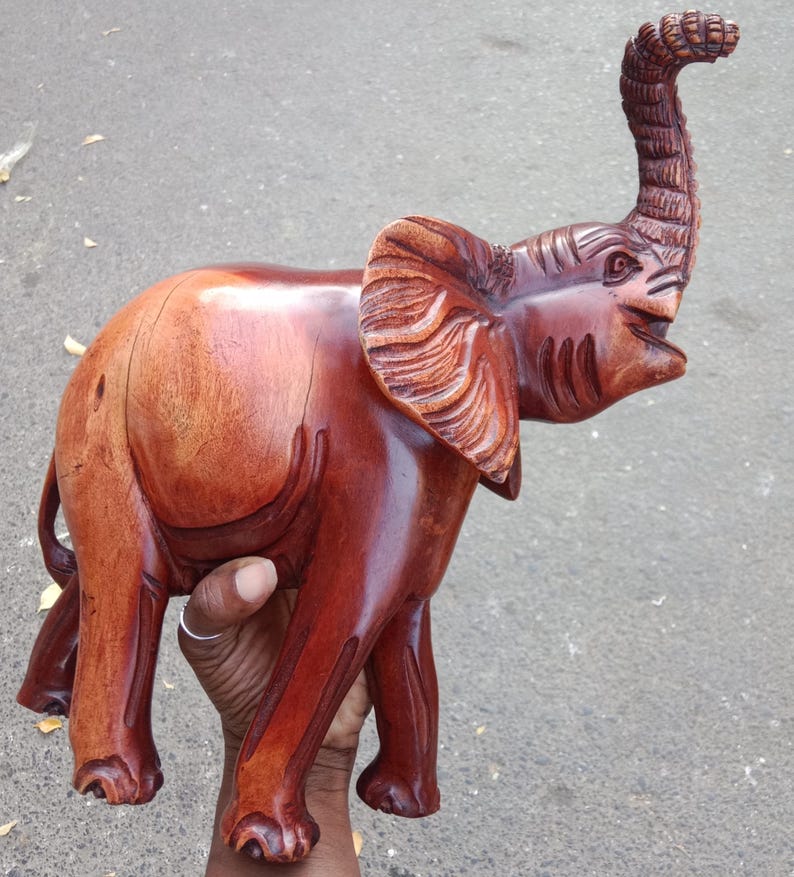 May include: A handcrafted wooden elephant sculpture with a polished, reddish-brown finish. The elephant has detailed carvings on its ears, trunk, and body, and is posed with its trunk raised. This decorative piece is a unique home decor item.