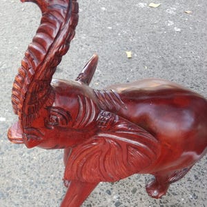 May include: A handcrafted, reddish-brown wooden elephant sculpture with intricate carved details. The elephant has a raised trunk, large ears, and a smooth, polished body. The sculpture is standing on four legs.