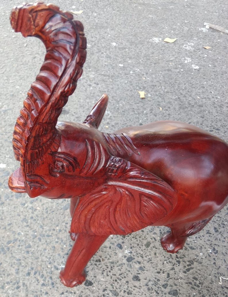 May include: A handcrafted, reddish-brown wooden elephant sculpture with intricate carved details. The elephant has a raised trunk, large ears, and a smooth, polished body. The sculpture is standing on four legs.