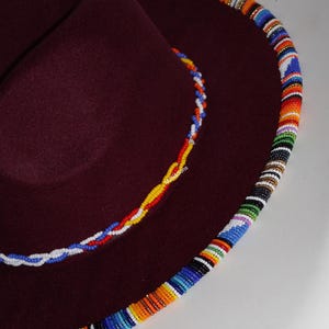 May include: A burgundy fedora hat with a colorful beaded trim around the brim. The hat features a smaller beaded accent on the crown, showcasing a vibrant mix of colors. The hat's design is a fusion of classic style and cultural artistry.