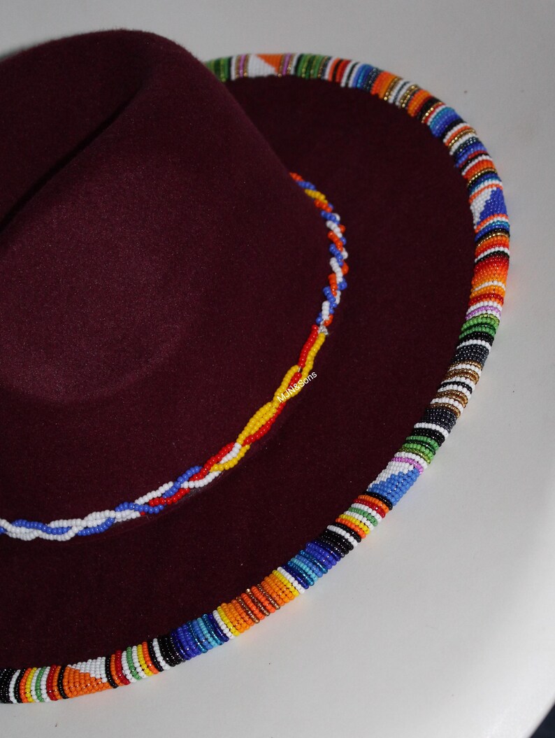 May include: A burgundy fedora hat with a colorful beaded trim around the brim. The hat features a smaller beaded accent on the crown, showcasing a vibrant mix of colors. The hat's design is a fusion of classic style and cultural artistry.