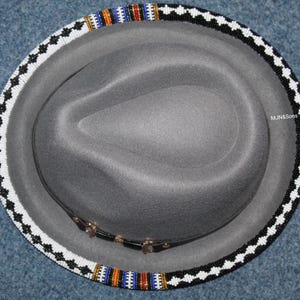 May include: A gray fedora hat with a beaded brim. The brim is decorated with black, white, orange, blue, and red beads. The hatband is black leather with gold-colored accents. The hat is a classic style.