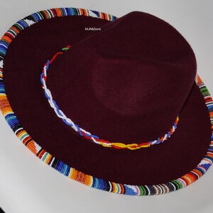 May include: A burgundy-colored hat with a wide brim and a beaded trim. The brim is adorned with a colorful, patterned beadwork in shades of blue, orange, and white. A smaller beaded band encircles the crown.
