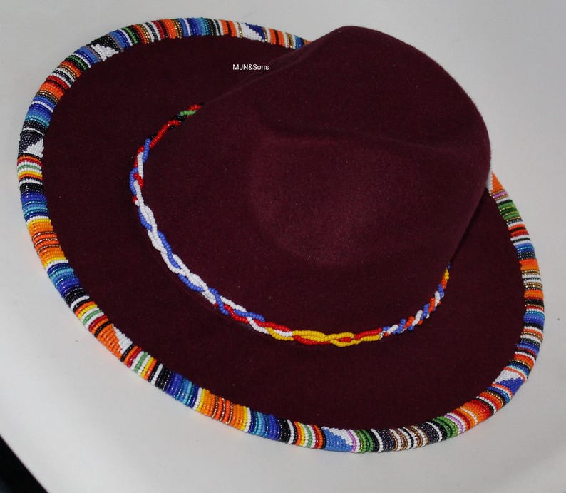 May include: A burgundy-colored hat with a wide brim and a beaded trim. The brim is adorned with a colorful, patterned beadwork in shades of blue, orange, and white. A smaller beaded band encircles the crown.