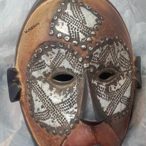 May include: A carved wooden mask with a brown, red, and white color scheme. The mask features open eye and mouth holes, with decorative silver-colored metal accents. The text "MJN&Sons" is visible on the left side.