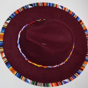 May include: A burgundy fedora-style hat with a wide brim. The brim and the crown are trimmed with colorful beaded patterns. The hat has a circular beaded design around the crown. The hat is a stylish accessory.