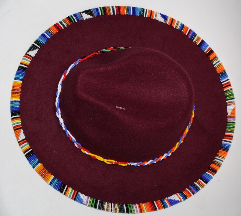 May include: A burgundy fedora-style hat with a wide brim. The brim and the crown are trimmed with colorful beaded patterns. The hat has a circular beaded design around the crown. The hat is a stylish accessory.