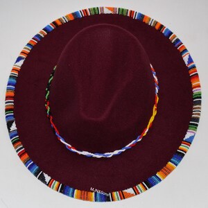 May include: A burgundy fedora-style hat with a wide brim. The brim is adorned with a colorful beaded trim. A smaller beaded band encircles the crown. The hat has the text "MJN&Sons" on the brim.